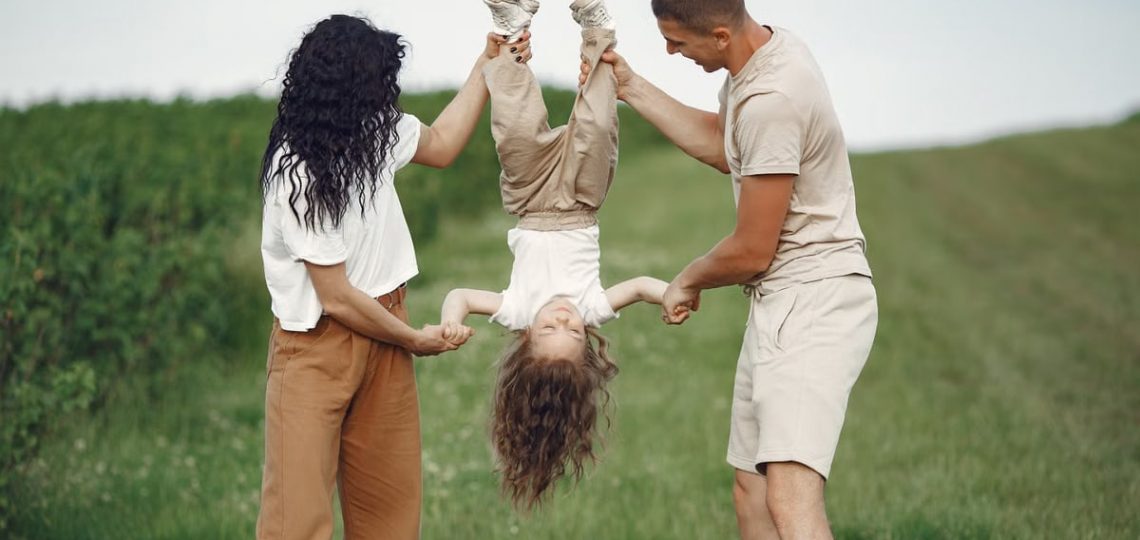 A joyful family enjoys outdoor play, lifting their child upside down in a green field.