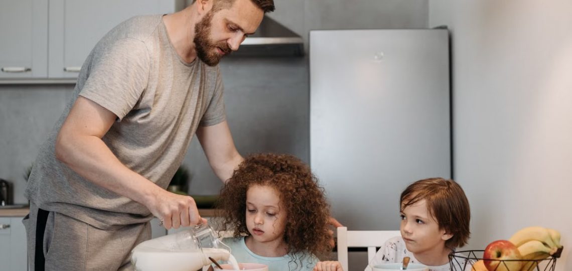 A father pours milk into cereal bowls for his children at the breakfast table in a modern kitchen.